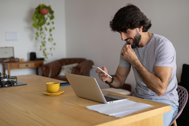 Photo un jeune homme travaille à la maison dans sa cuisine avec un ordinateur portable et des papiers sur le bureau en bois de la cuisine carnet de notes gris