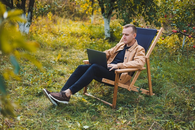 Un jeune homme travaille sur un ordinateur portable à l'extérieur Un jeune pigiste se repose dans la forêt Travail à distance loisirs actifs en été Tourisme concept de personnes homme assis sur une chaise à l'extérieur