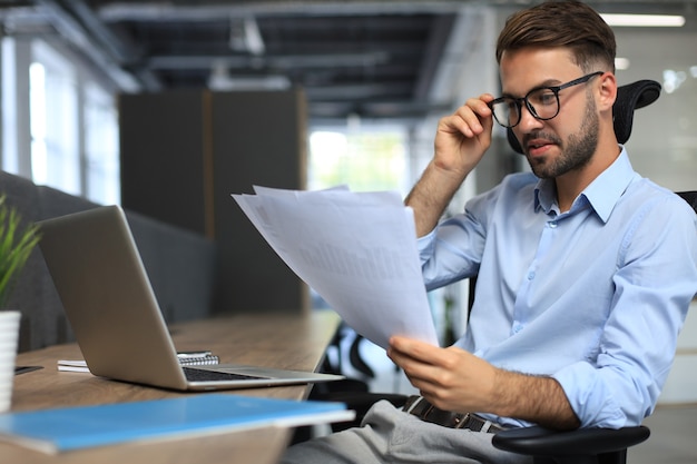 Un jeune homme travaille avec des papiers alors qu'il est assis au bureau. L'entrepreneur à succès étudie les documents avec un regard attentif et concentré.