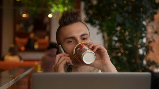 Un jeune homme travaille sur son ordinateur portable dans un café et parle au téléphone.