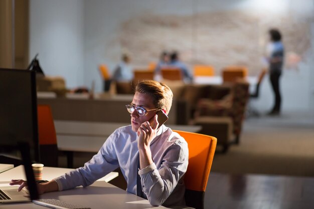 Jeune homme utilisant un téléphone portable tout en travaillant sur ordinateur la nuit dans un bureau sombre.