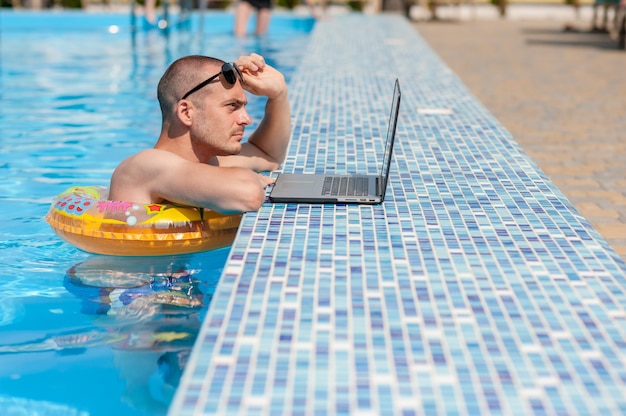 Le jeune homme en vacances d'été avec l'ordinateur portable, les affaires en ligne. Bureau à domicile à distance dans la piscine en anneau de caoutchouc en quarantaine