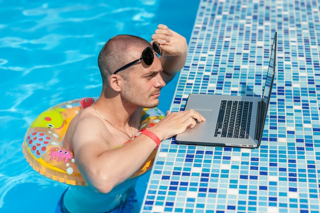 Le jeune homme en vacances d'été avec l'ordinateur portable, les affaires en ligne. Bureau à domicile à distance dans la piscine en anneau de caoutchouc en quarantaine