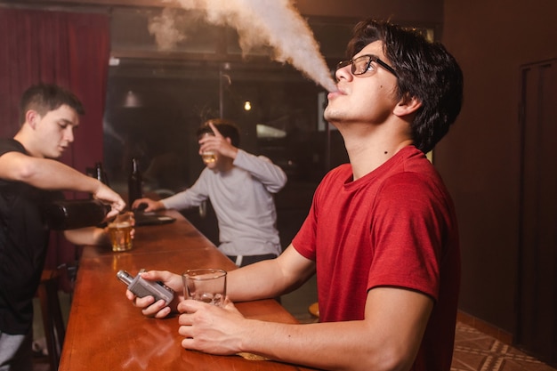 Un jeune homme vapotant pendant que ses amis boivent de la bière au bar.