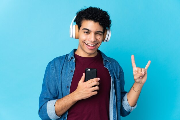 Jeune homme vénézuélien isolé sur bleu écoute de la musique avec un mobile faisant un geste rock