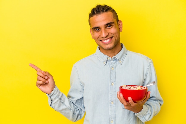 Jeune homme vénézuélien tenant un bol de céréales isolé sur fond jaune souriant et pointant de côté