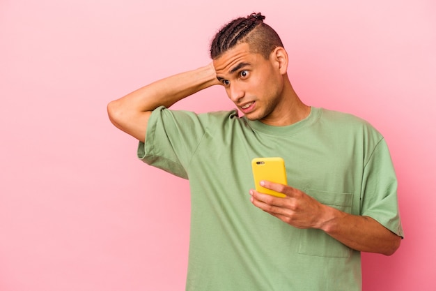 Jeune homme vénézuélien tenant un téléphone portable isolé sur fond rose touchant l'arrière de la tête, pensant et faisant un choix.