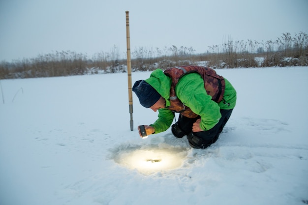 Jeune homme vérifie un trou dans un lac gelé sur un pêcheur d'hiver