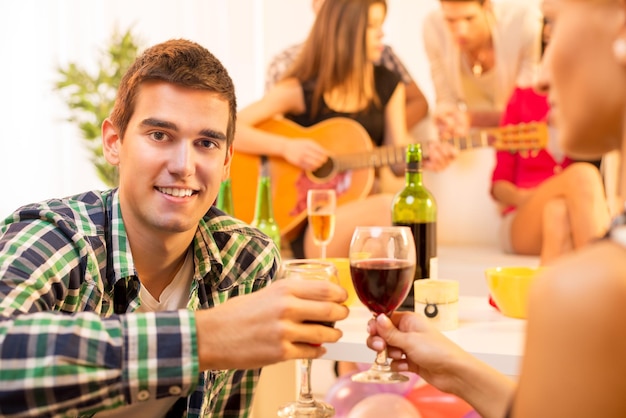 Un jeune homme avec un verre de vin lors de la fête à la maison porte un toast avec une fille, et en arrière-plan, vous pouvez voir un groupe de jeunes réunis autour de la fille avec une guitare acoustique.