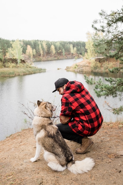 Jeune homme en veste à carreaux et jeans noirs assis sur des squats par mignon animal de race pure tout en se refroidissant au bord du lac dans la forêt