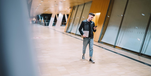 jeune homme en veste noire et denim bleu utilisant un téléphone portable et tenant un ordinateur portable en marchant