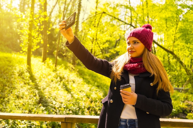 Jeune homme avec une veste noire, une écharpe et un bonnet de laine rouge profitant d'un parc en automne, prenant un selfie avec le mobile