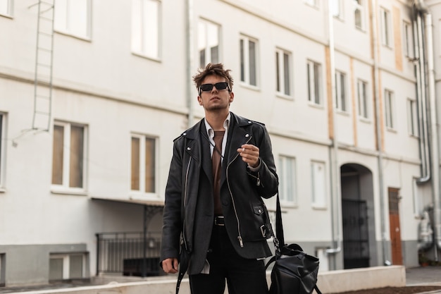 Jeune homme en vêtements de cuir noir surdimensionnés pour les jeunes de la mode avec support de sac à dos avec cigarette près des bâtiments de la ville un jour de printemps. Un hipster brutal dans des lunettes de soleil sombres profite d'un tabagisme à l'extérieur
