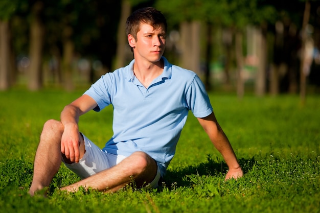 Jeune homme en vêtements décontractés assis sur l'herbe verte et regardant de côté dans le parc par temps clair d'été. Liberté intérieure et concept de mode de vie heureux