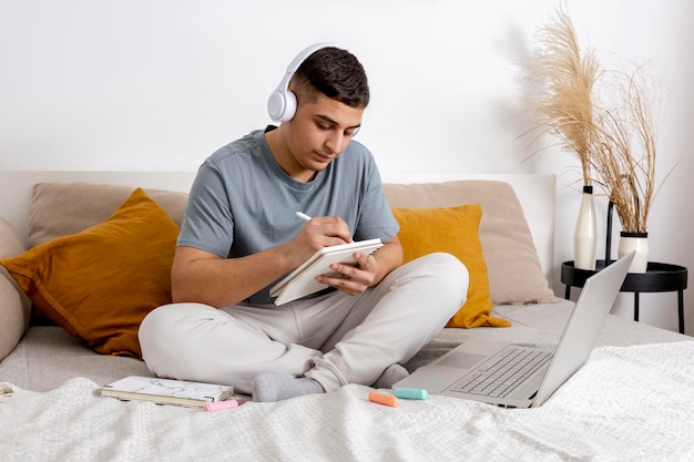 Jeune homme avec des vêtements décontractés assis sur le lit à la maison avec un ordinateur portable et étudiant Homme utilisant une plateforme d'apprentissage en ligne pour passer un appel vidéo avec son professeur Enseignement à distance Intérieur confortable