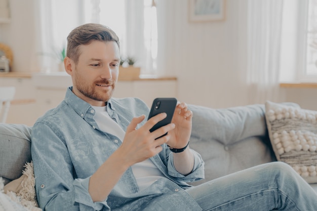 Jeune homme en vêtements décontractés surfant sur le Web sur son téléphone et se relaxant, vérifiant les messages et les notifications tout en étant assis sur un canapé confortable avec les jambes croisées, arrière-plan flou