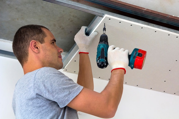 Jeune homme en vêtements et gants de travail habituels fixant le plafond suspendu de cloisons sèches au cadre métallique à l'aide d'un tournevis électrique au plafond isolé avec du papier d'aluminium brillant. DIY, faites-le vous-même concept.