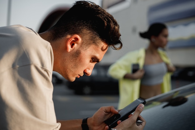 Un jeune homme vêtu d'un t-shirt blanc utilise un téléphone à côté de la fille sur la place pour se garer par beau temps.