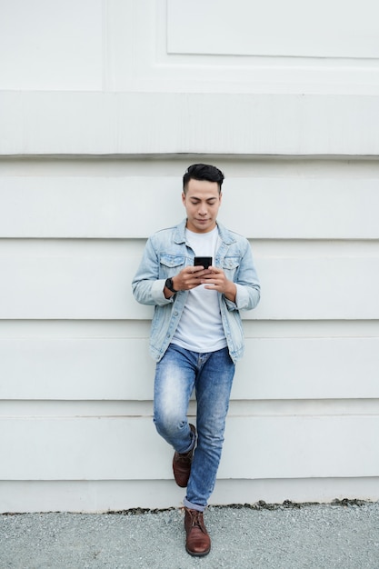 Jeune homme vietnamien à la mode sérieux dans des vêtements de denim tenant le pied sur le mur et utilisant le téléphone moderne