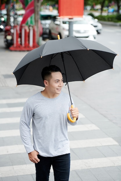 Jeune homme vietnamien positif en pull gris marchant avec un parapluie noir sur le passage pour piétons en ville