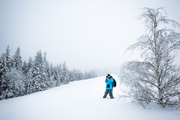 Jeune homme voyageur avec sac à dos prend des photos de beau grand sapin enneigé dans une neige élevée sur fond de brouillard sur une journée d'hiver glaciale