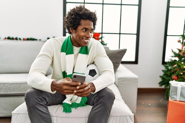 Jeune hooligan afro-américain utilisant un smartphone soutenant l'équipe de football assis sur le canapé à la maison.