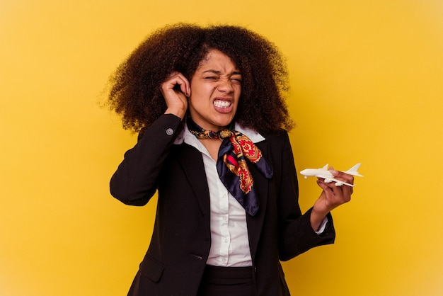 Jeune hôtesse de l'air afro-américaine tenant un petit avion isolé sur fond jaune couvrant les oreilles avec les mains.