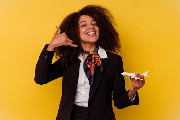 Jeune hôtesse de l'air afro-américaine tenant un petit avion isolé sur un mur jaune montrant un geste d'appel de téléphone mobile avec les doigts.