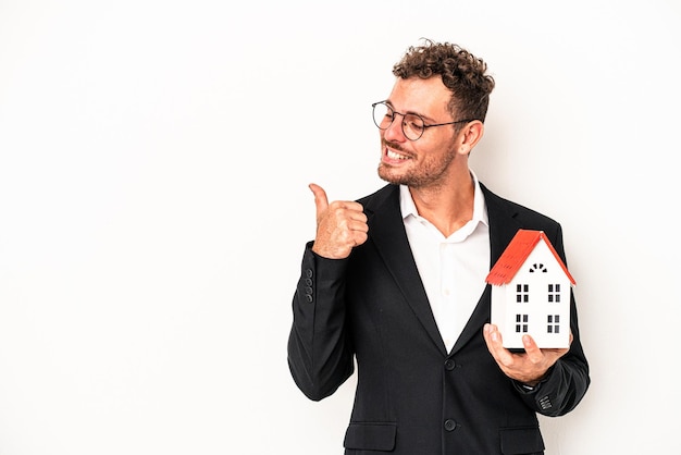 Jeune immobilier caucasien tenant une maison de jouets isolée sur fond blanc avec le pouce loin, riant et insouciant.