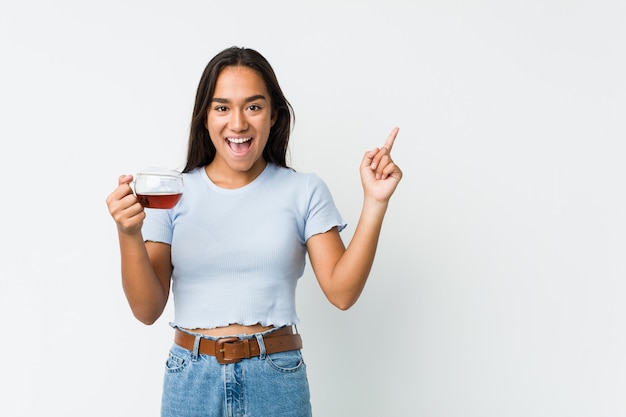 Jeune Indien de race mixte tenant une tasse de thé souriant joyeusement pointant avec l'index loin.