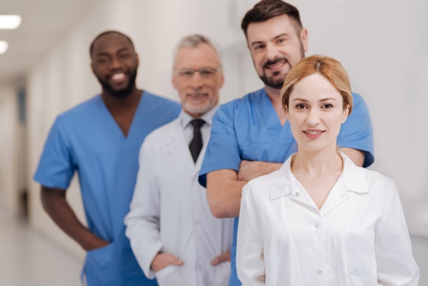 Jeune infirmière charmante joyeuse bénéficiant de responsabilités au travail et debout à l'hôpital tandis que ses collègues debout