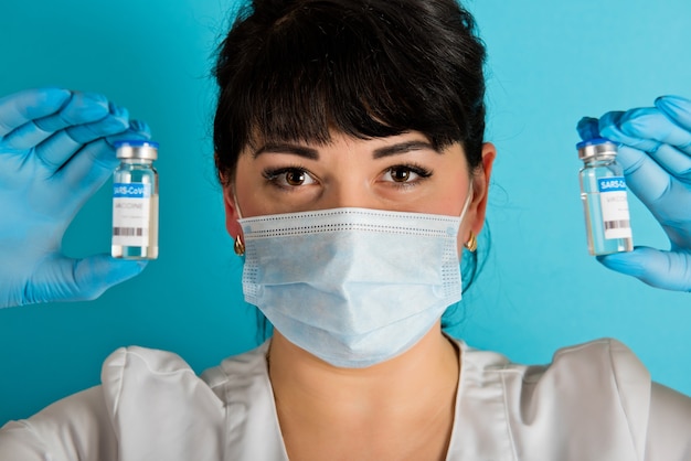 Jeune infirmière dans un masque médical tenant deux flacons de vaccin contre le coronavirus Covid-19 sur fond bleu. Fermer.