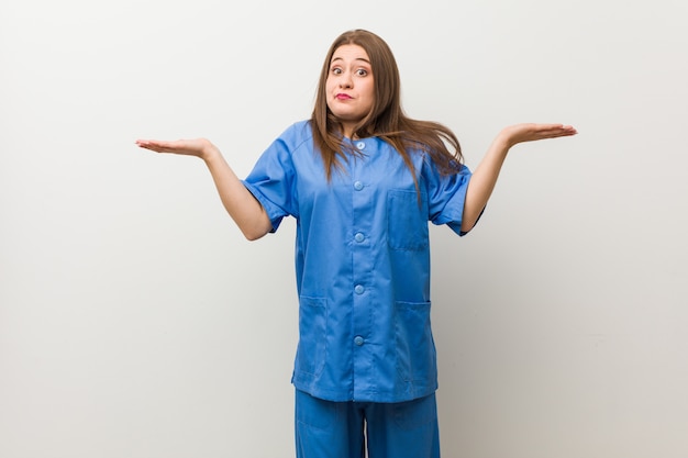 Jeune infirmière femme contre un mur blanc haussant les épaules de manière confuse et douteuse de tenir un espace de copie.