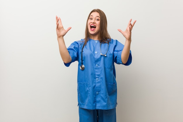 Jeune infirmière femme contre un mur blanc recevant une agréable surprise, excitée et levant les mains