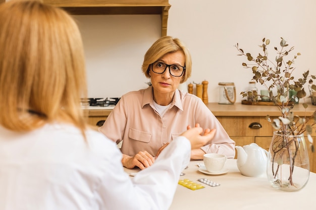 Une jeune infirmière fournit un service médical de soins aide à soutenir une femme mûre âgée lors d'une visite médicale à domicile, une femme médecin soignant donne de l'empathie pour encourager le patient retraité dans la cuisine.