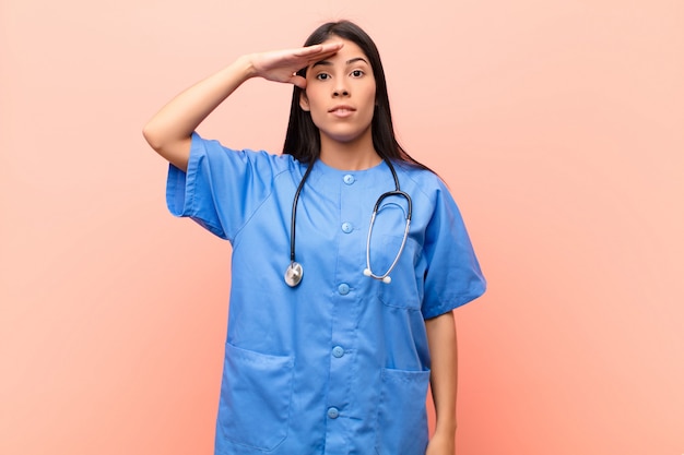 Jeune infirmière latine saluant avec un salut militaire dans un acte d'honneur et de patriotisme, montrant du respect contre le mur rose