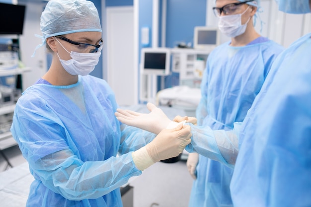 Jeune infirmière en masque de protection et vêtements de travail aidant le chirurgien avec des gants tout en se préparant à l'opération