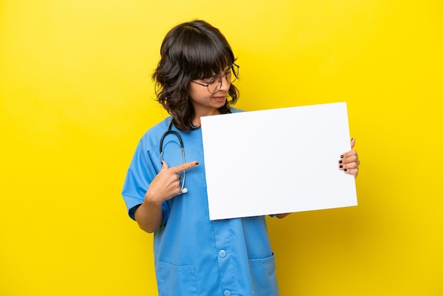 Jeune infirmière médecin femme isolée sur fond jaune tenant une pancarte vide avec une expression heureuse et la pointant
