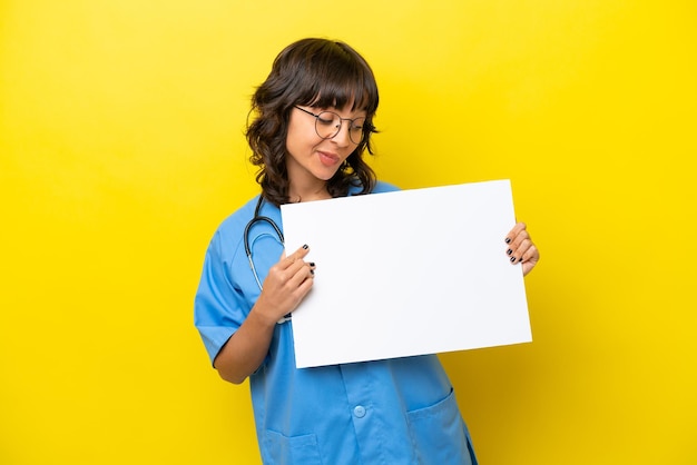 Jeune infirmière médecin femme isolée sur fond jaune tenant une pancarte vide avec une expression heureuse