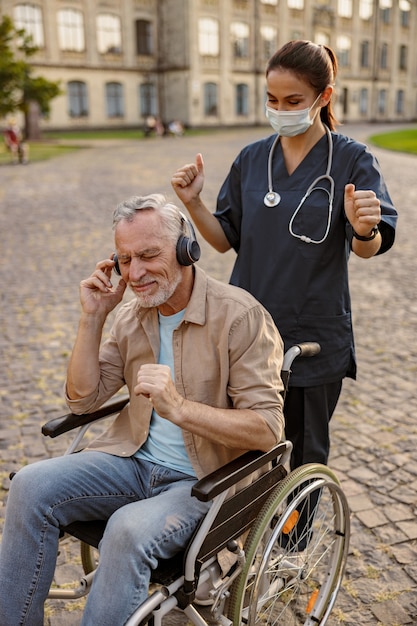 Jeune infirmière portant un masque de protection prenant soin d'un patient âgé en convalescence en fauteuil roulant