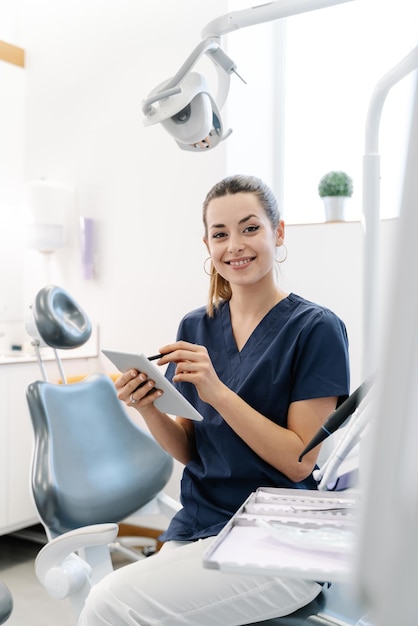 Une jeune infirmière portant une veste bleue sourit dans le cabinet dentaire avant de recevoir le patient et manipule une tablette