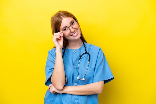 Jeune infirmière rousse femme isolée sur fond jaune avec des lunettes et heureux