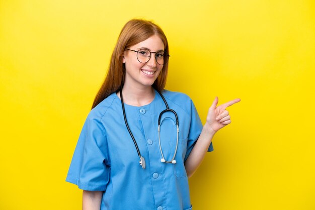 Jeune infirmière rousse femme isolée sur fond jaune pointant le doigt sur le côté