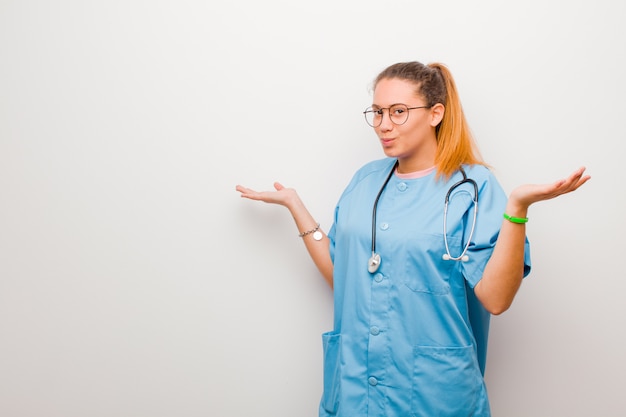 Photo jeune infirmière se sentant perplexe et confuse, doutant, pondérant ou choisissant différentes options avec une drôle d'expression contre le mur blanc