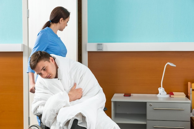 Jeune infirmière en uniforme tirant un fauteuil roulant avec un patient malade recouvert d'une couette dans une salle d'hôpital. Concept de soins de santé
