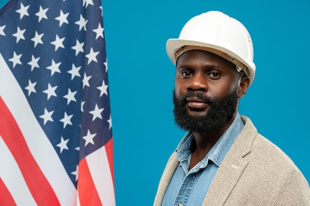 Jeune ingénieur afro-américain barbu en casque de protection et tenues de soirée debout contre le drapeau américain et bleu