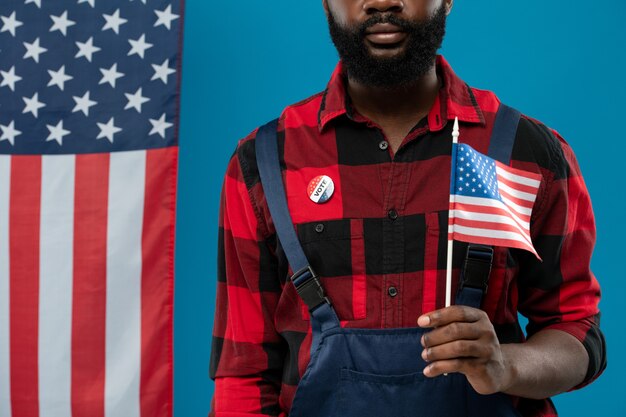 Jeune ingénieur afro-américain barbu en tenue de travail tenant un drapeau contre le bleu avec des étoiles et des rayures sur la droite