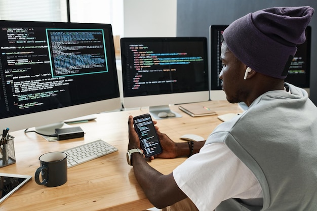 Jeune ingénieur informatique sérieux dans les écouteurs faisant défiler les données codées dans le téléphone mobile tout en étant assis près