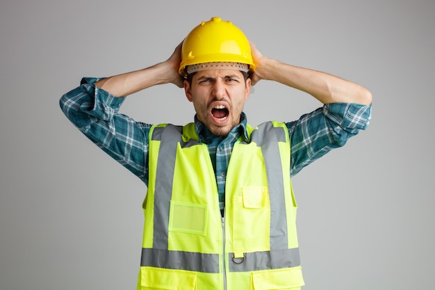 Jeune ingénieur masculin stressé portant un casque de sécurité et un uniforme regardant la caméra en gardant les mains sur la tête en criant isolé sur fond blanc