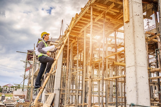 Jeune ingénieur professionnel en casque de protection et papier de plans sur place travaillant sur une échelle sur le chantier de construction de la maison